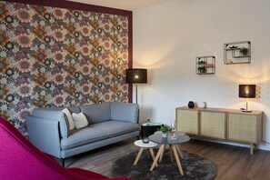 A living room with a floral wallpaper, a grey sofa, a round coffee table, a cabinet with plants, and two wall-mounted lamps. - Nautimar Apartment Kattegat 32 (Büsum)