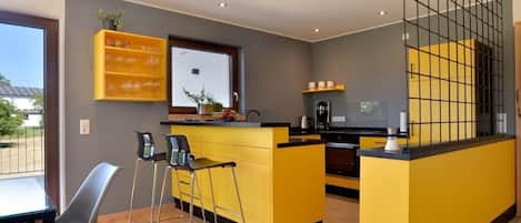Une cuisine moderne avec un îlot central jaune, des tabourets de bar et une vue à travers une porte vitrée.