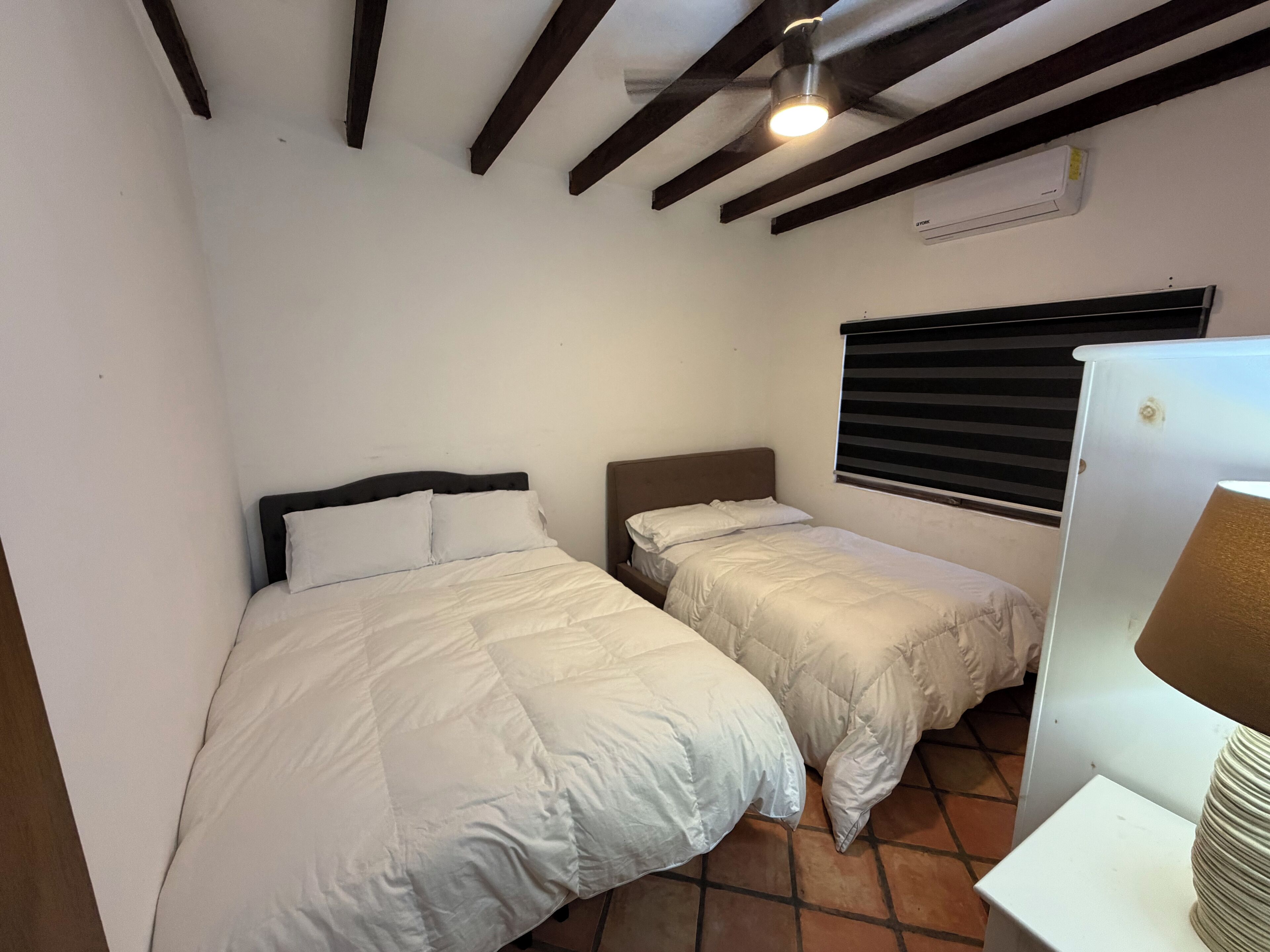 Quarto pequeno e compacto com duas camas, ventilador de teto, ar condicionado e janela com persianas.