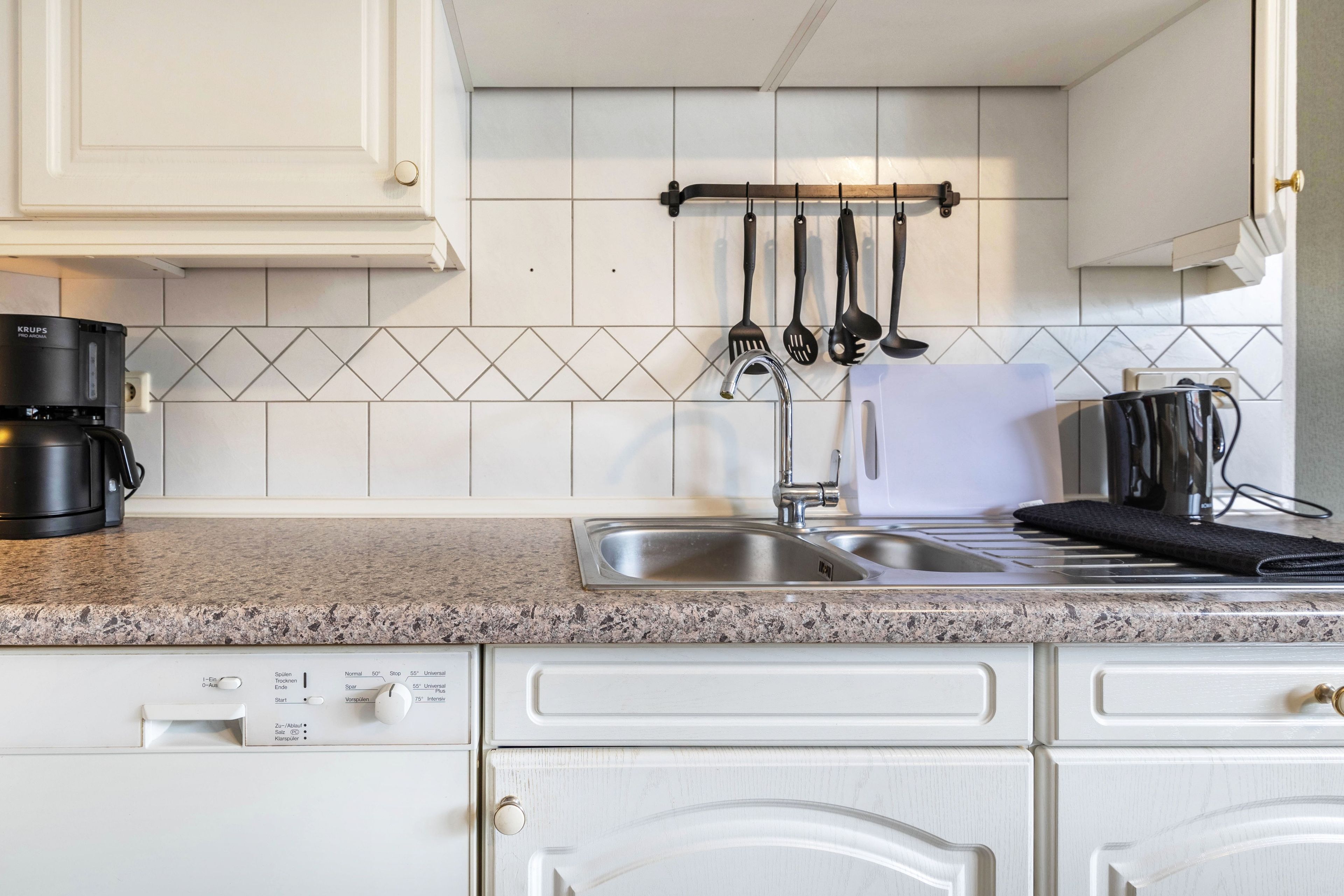 Una cocina con gabinetes blancos, encimera de granito, cafetera, pileta y utensilios colgantes.