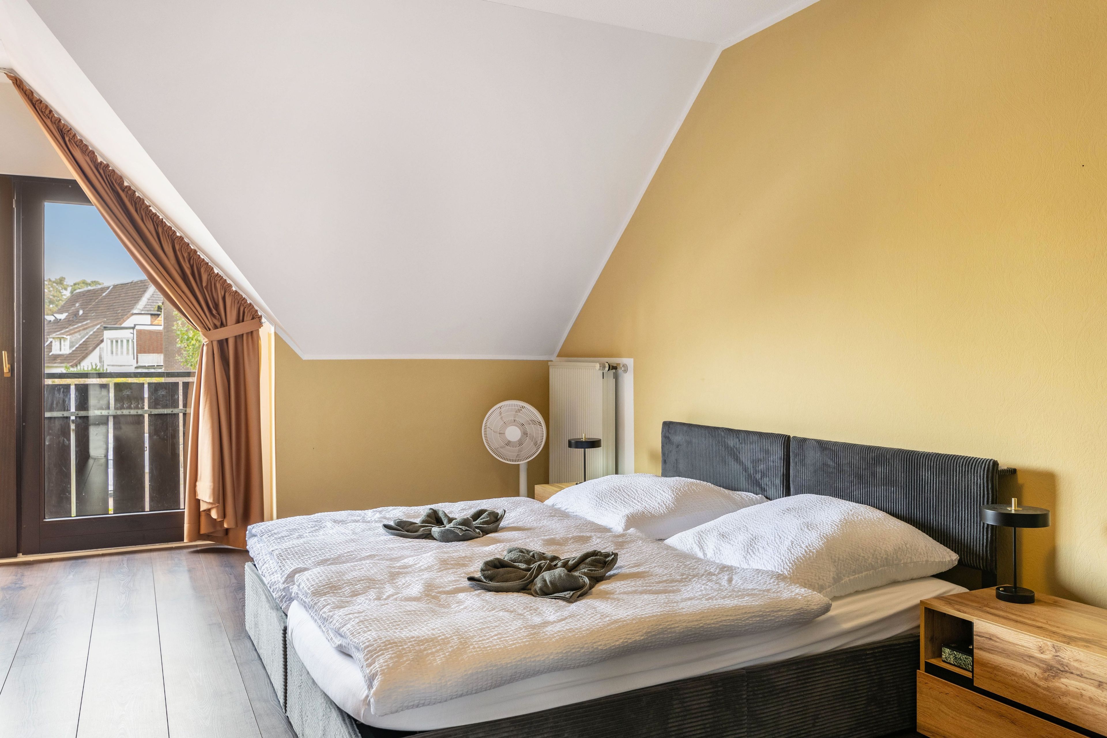 A bedroom with a sloped ceiling, a bed with a white duvet, a bedside table with a lamp, and a view of the outside through a window with curtains.