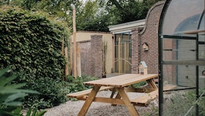 A wooden picnic table in a garden with a brick wall and a glass greenhouse structure.