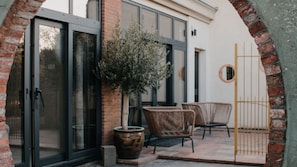 A patio area with a brick wall, a potted plant, wicker furniture, and a metal gate. - Olijf Room | Double Room with Private Bathroom | Near Utrecht (Maarssen)