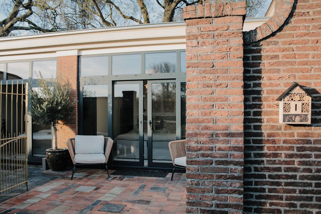 Un bâtiment moderne doté d’une entrée avec une porte en verre, d’un espace extérieur aménagé pour s’asseoir et d’un mur en briques orné d’une nichée décorative pour oiseaux.