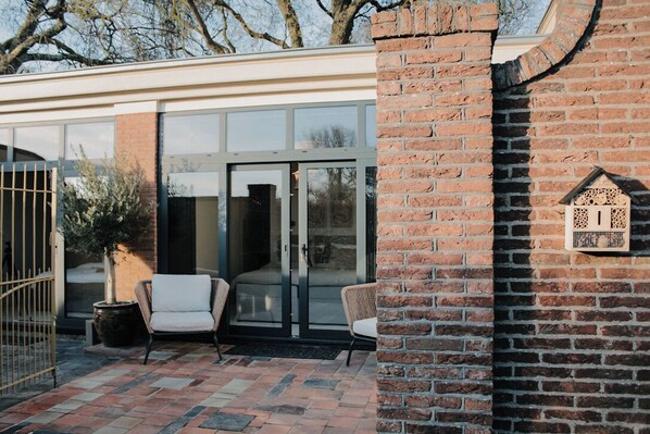 Un bâtiment moderne doté d’une entrée avec une porte en verre, d’un espace extérieur aménagé pour s’asseoir et d’un mur en briques orné d’une nichée décorative pour oiseaux.