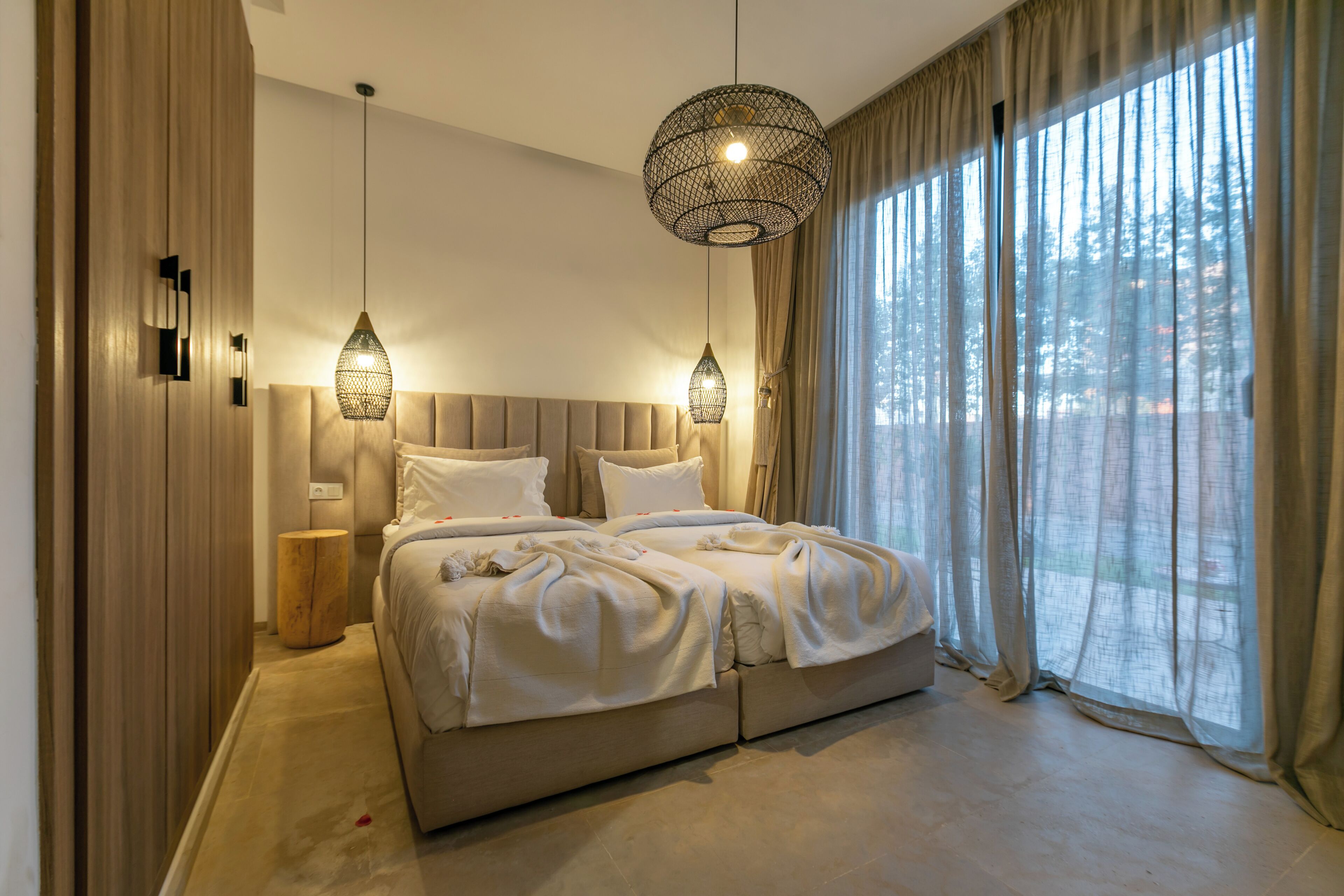 Une chambre à coucher avec un grand lit, deux tables de chevet, un luminaire suspendu et une vue sur l’extérieur à travers une fenêtre avec des rideaux.