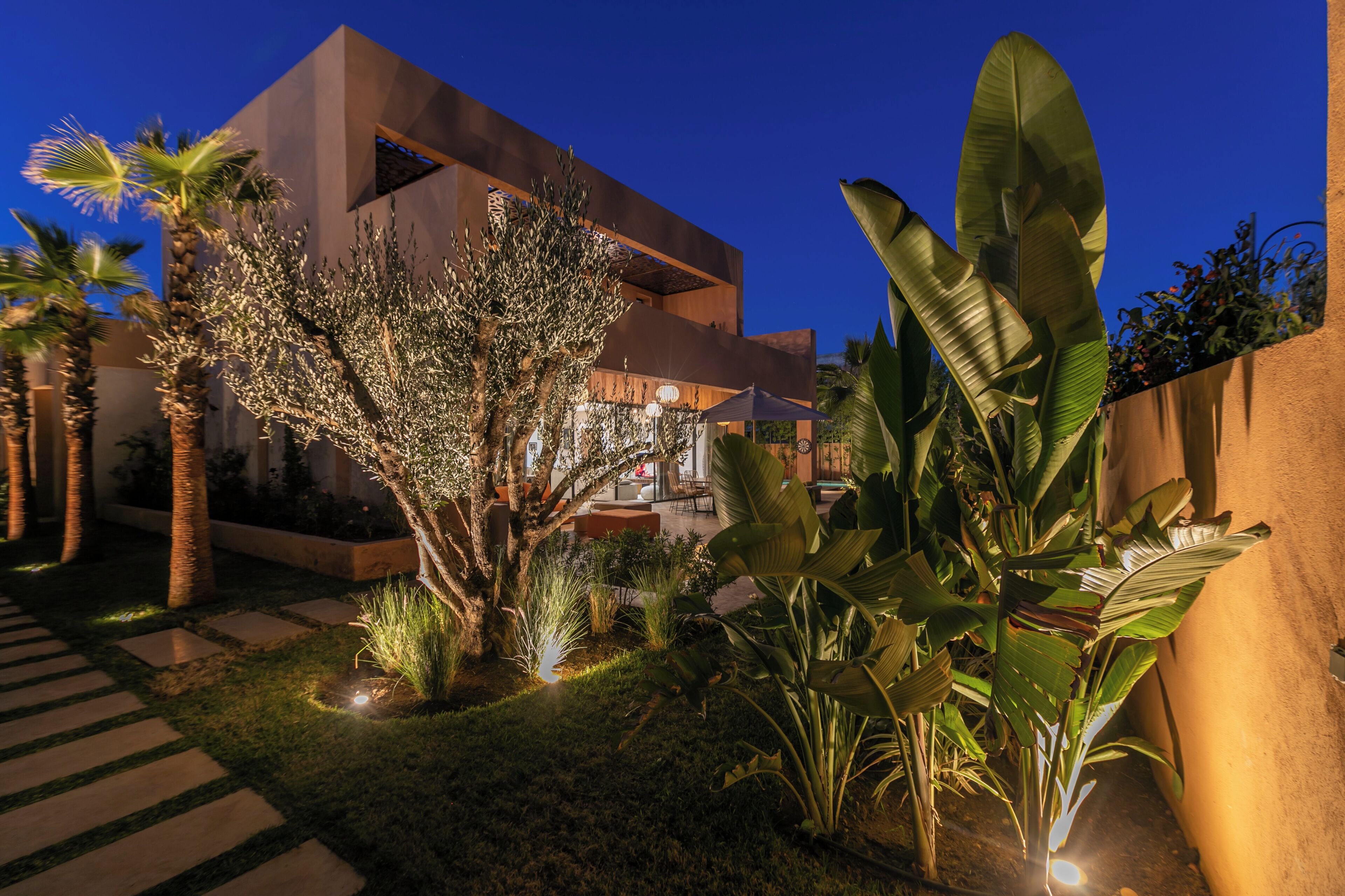 Un bâtiment moderne à la façade géométrique, entouré d’un jardin bien entretenu où poussent différentes espèces de plantes, et agrémenté d’un chemin pavé.
