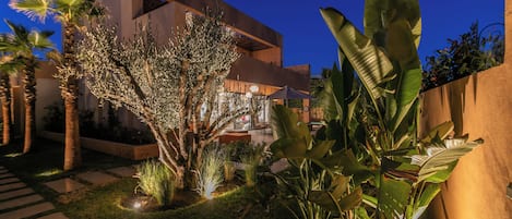 Un bâtiment moderne à la façade géométrique, entouré d’un jardin bien entretenu où poussent différentes espèces de plantes, et agrémenté d’un chemin pavé.