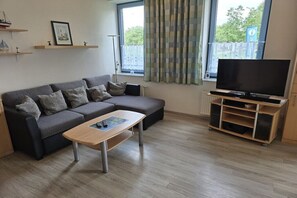 Una sala de estar moderna con un sofá gris, una mesa de centro de madera, un televisor de pantalla plana sobre un mueble de madera y una ventana con cortinas.