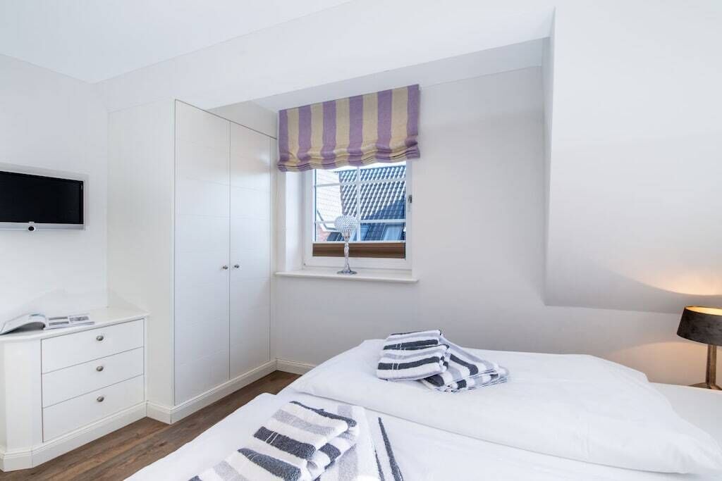 A bedroom with a bed, a bedside table, a TV mounted on the wall, and a window with a striped valance.
