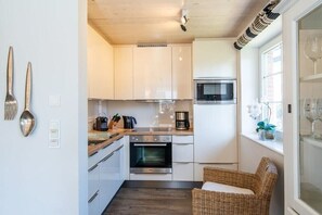 A compact kitchen with white cabinets, a microwave, an oven, and a wicker chair by the window. - Holiday home in Sylt with 3 bedrooms (Westerland)
