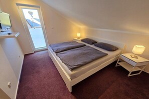 A bedroom with a bed, bedside lamps, a TV on a shelf, and a door leading to an outdoor area. - Pure Holiday Castle 007 (Burg auf Fehmarn)
