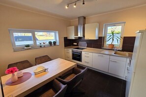A modern kitchen with a dining area, featuring a central island, built-in cabinets, and a window with plants. - Pure Holiday Castle 007 (Burg auf Fehmarn)