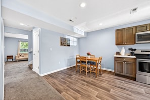 A modern kitchen with wooden cabinets, a dining area with a table and chairs, and a living room with a sofa and a window. - Roadmap Lodging: Central J, 2BR Downtown Apartment, Monthly (Columbus)