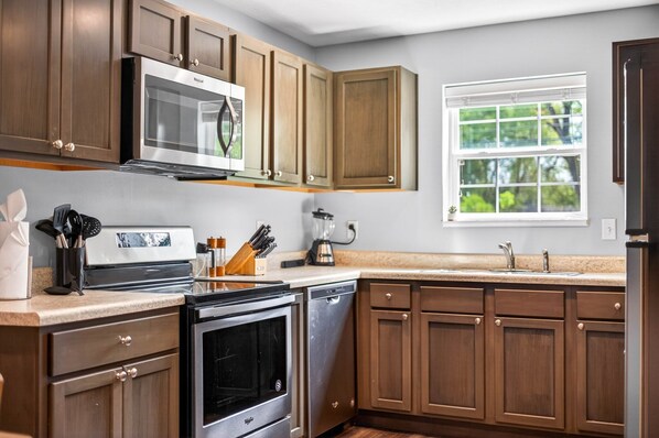 A kitchen with wooden cabinets, stainless steel appliances, a window with a view of greenery, and a countertop with various kitchen items.