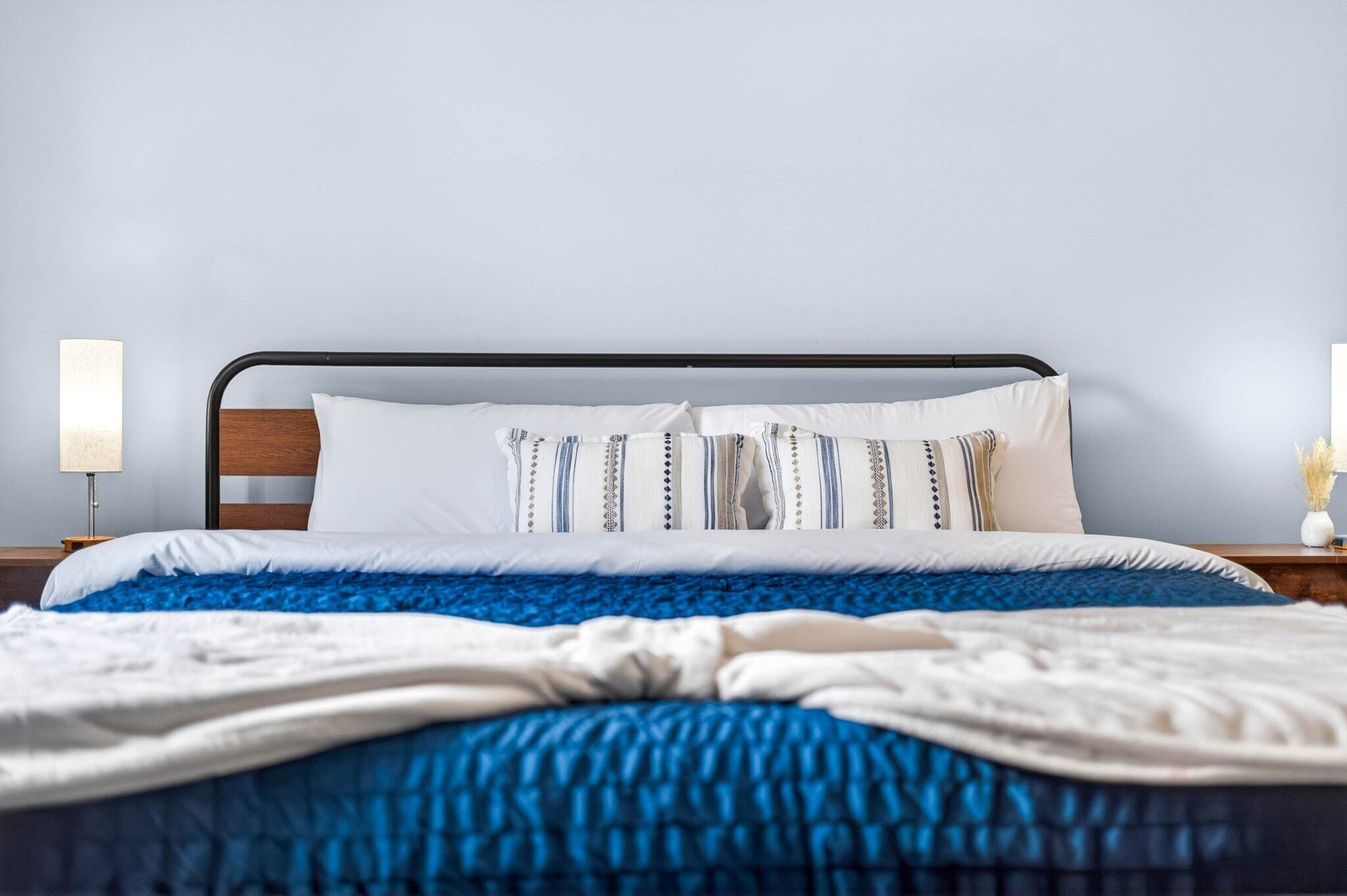Ein ordentlich bezogenes Bett mit blauer Decke und weißer Bettwäsche, einem schwarzen Metall-Kopfteil und zwei Nachttischlampen auf Holznachttischen.
