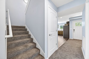 A staircase with carpeted steps leading to a door, a kitchen area with cabinets and a refrigerator, and a hallway with carpet flooring. - Roadmap Lodging: Central J, 2BR Downtown Apartment, Monthly (Columbus)