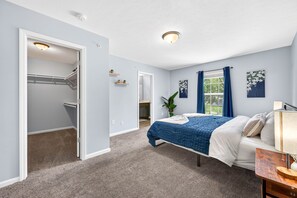 Un dormitorio con cama, mesita de noche, ventana con cortinas, armario y puerta que da a otra habitación.