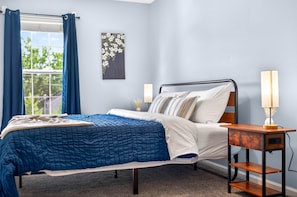 A bedroom with a bed, a nightstand, a lamp, and a window with curtains. - Roadmap Lodging: Central J, 2BR Downtown Apartment, Monthly (Columbus)