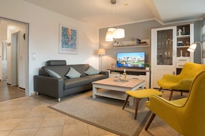 A modern living room with a sofa, coffee table, and yellow chairs. There is a television on a stand and a cabinet with glass doors. - Holiday apartment with 1 bedroom (Greetsiel)