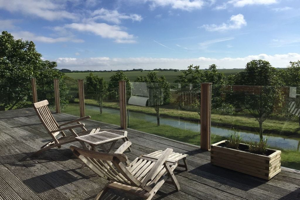 A wooden deck with two lounge chairs and a planter box, overlooking a body of water and a grassy landscape.