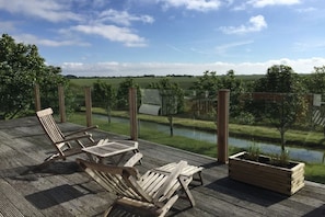 Eine Holzterrasse mit zwei Liegestühlen und einem Blumenkasten, mit Blick auf ein Gewässer und eine grüne Landschaft.
