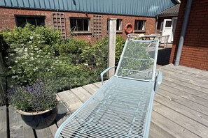 Une chaise longue en métal sur une terrasse en bois, avec un bâtiment en briques en arrière-plan.