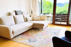 A living room with a beige sectional sofa, a colorful rug, and a view of greenery through large windows. - Floor cutting suites "Wanderlust" (Tegernsee)