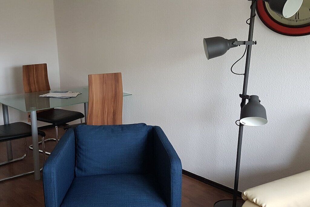 Une pièce avec un fauteuil bleu, une table en verre entourée de chaises, un lampadaire et une horloge au mur.