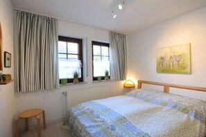 Une chambre à coucher avec un lit, deux fenêtres avec des rideaux, une lampe de chevet, un petit tabouret et un tableau au mur.