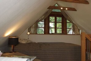 Ein gemütliches Schlafzimmer unter dem Dach mit geneigter Decke, einem großen Fenster mit Vorhängen, einem Bett mit gestreifter Decke und einer Nachttischlampe.
