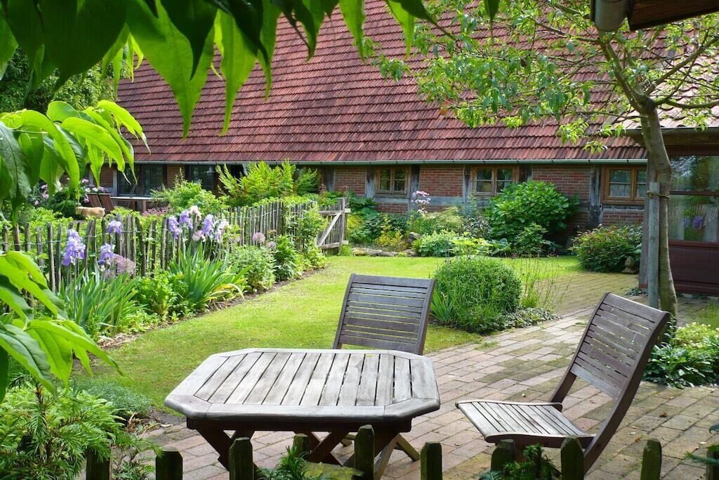 Un jardín con una mesa de madera y dos sillas, una casa de ladrillo con techo rojo, y diversas plantas y flores.