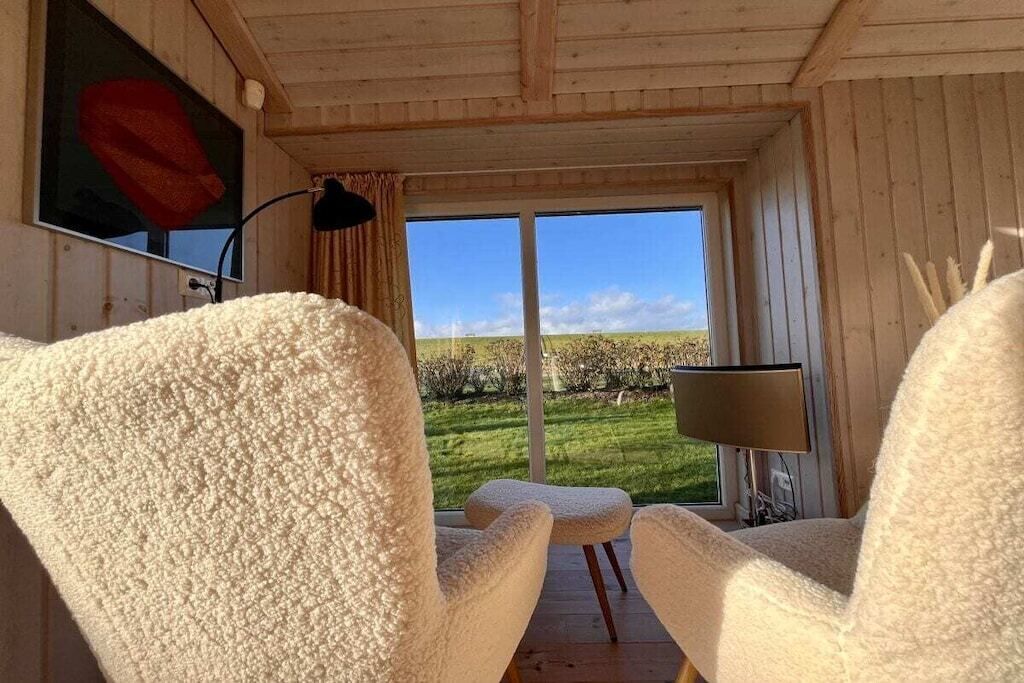 Una habitación con techo de madera, dos sillas blancas, una mesa, una lámpara y un ventanal con vistas a un paisaje verde.