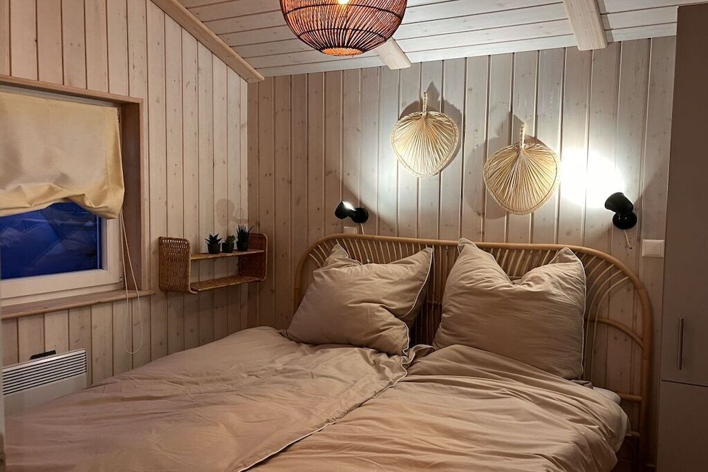 Un dormitorio con pared de paneles de madera, cama con ropa de cama color beige, dos lámparas de pared y una ventana con vistas.