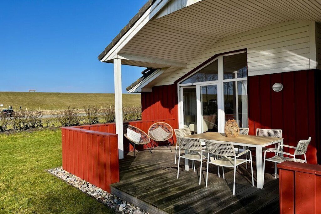 Un patio techado al aire libre con mesa y sillas de madera, una pared roja y vista a un campo cubierto de césped.