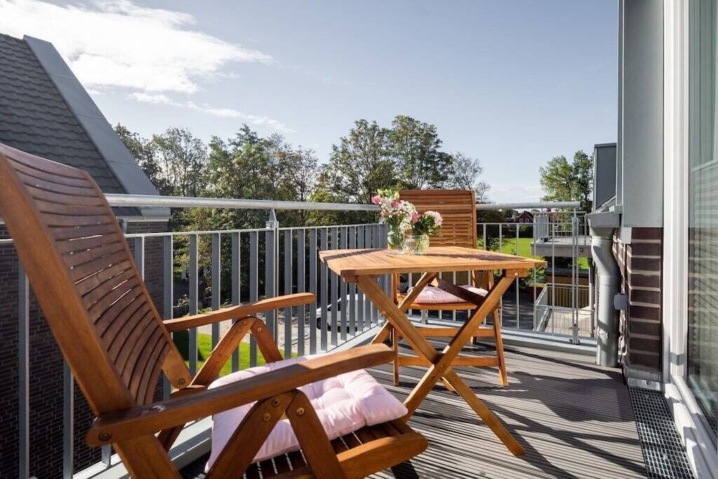 A balcony with a wooden chair and table, a flower arrangement, and a view of trees and buildings.