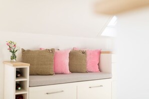 Ein Zimmer mit einer Bank, Kissen, einem kleinen Regal mit Flaschen und einer Vase mit Blumen.