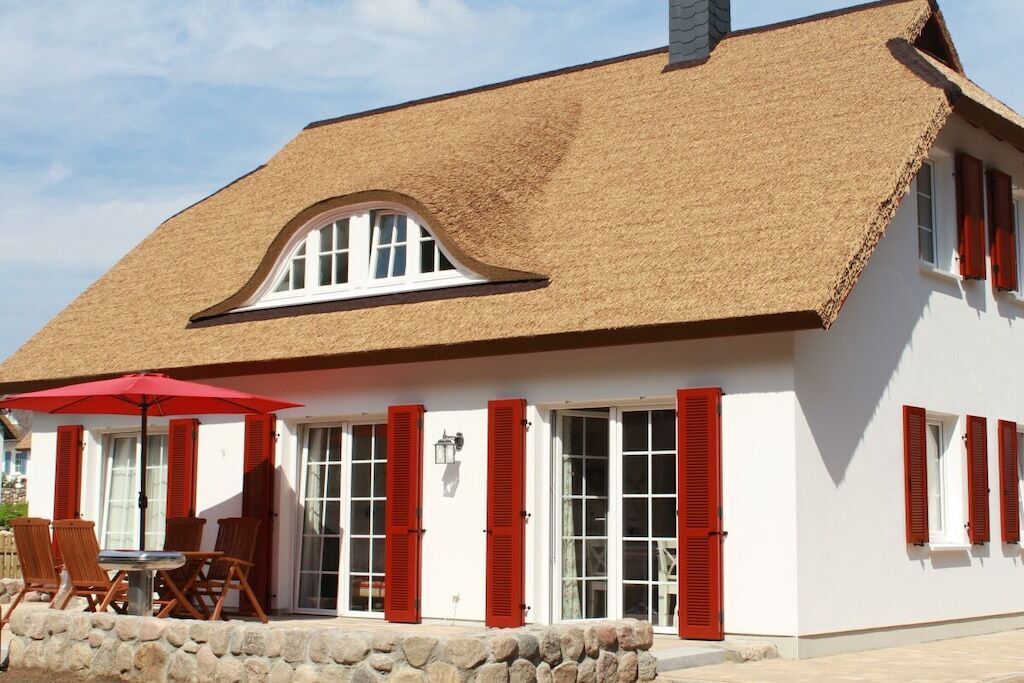 A building with a thatched roof, white walls, and red shutters. There is a table with chairs and a red umbrella outside.