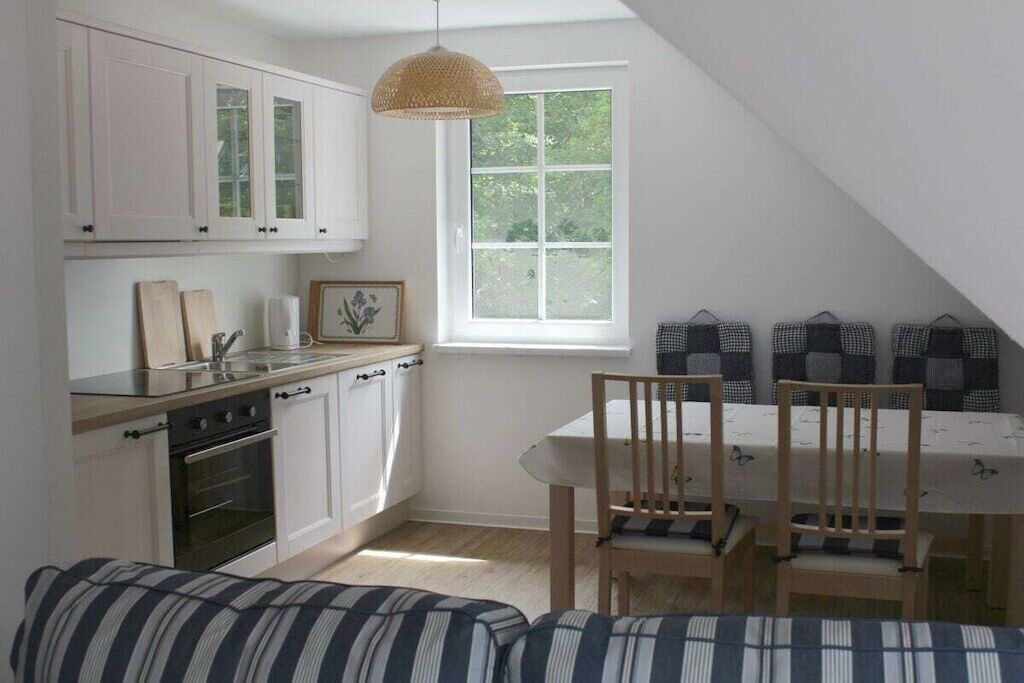 Et lille køkken med hvide skabe, et træspisebord med stole og et vindue med udsigt over grønne områder.