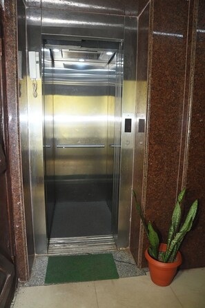Un ascensor con pantalla digital, un felpudo verde en la entrada y una planta en maceta a su lado.