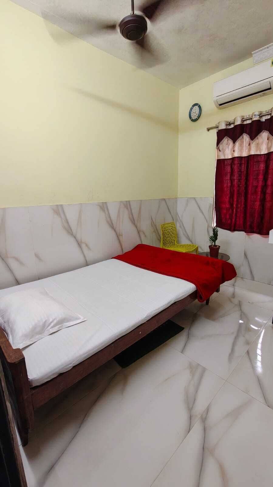 Uma cama de solteiro com estrutura de madeira, um cobertor vermelho e um travesseiro branco em um quarto com piso de mármore e ventilador de teto.