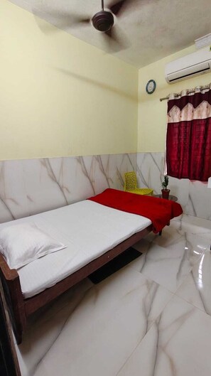 Una cama individual con estructura de madera, una colcha roja y un almohadón blanco en una habitación con suelo de mármol y ventilador de techo.