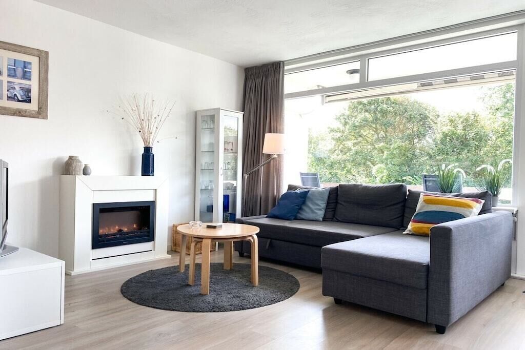 A modern living room with a fireplace, grey sofa, and a large window with a view of greenery.