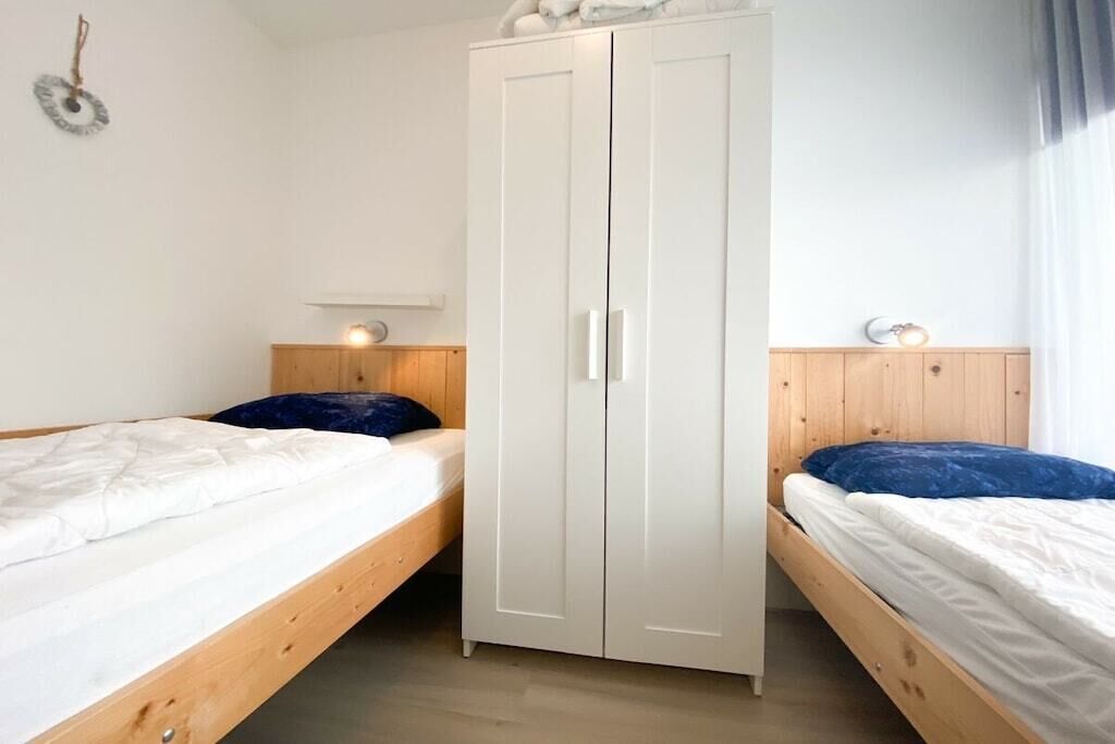 Quarto com duas camas de solteiro, guarda-roupa branco, cabeceiras de madeira e luminária fixada na parede.