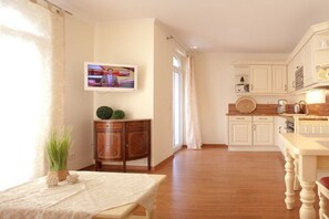 A well-lit room with a wooden floor, a dining table with a white tablecloth, a television mounted on the wall, and a kitchen area with white cabinets and a wooden countertop. - Villa Lena Apt. 7, Nagel Family (Sellin)