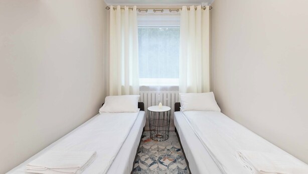 Een kleine, schone kamer met twee éénpersoonsbedden, een klein rond tafeltje en een raam met dunne gordijnen.