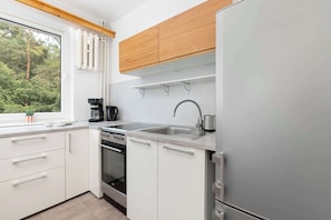 Una cocina moderna con armarios blancos, un refrigerador de acero inoxidable, horno empotrado y ventana con vista a la vegetación.