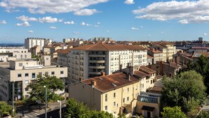 A cityscape with a mix of modern and traditional buildings, a clear sky, and a distant view of the sea.