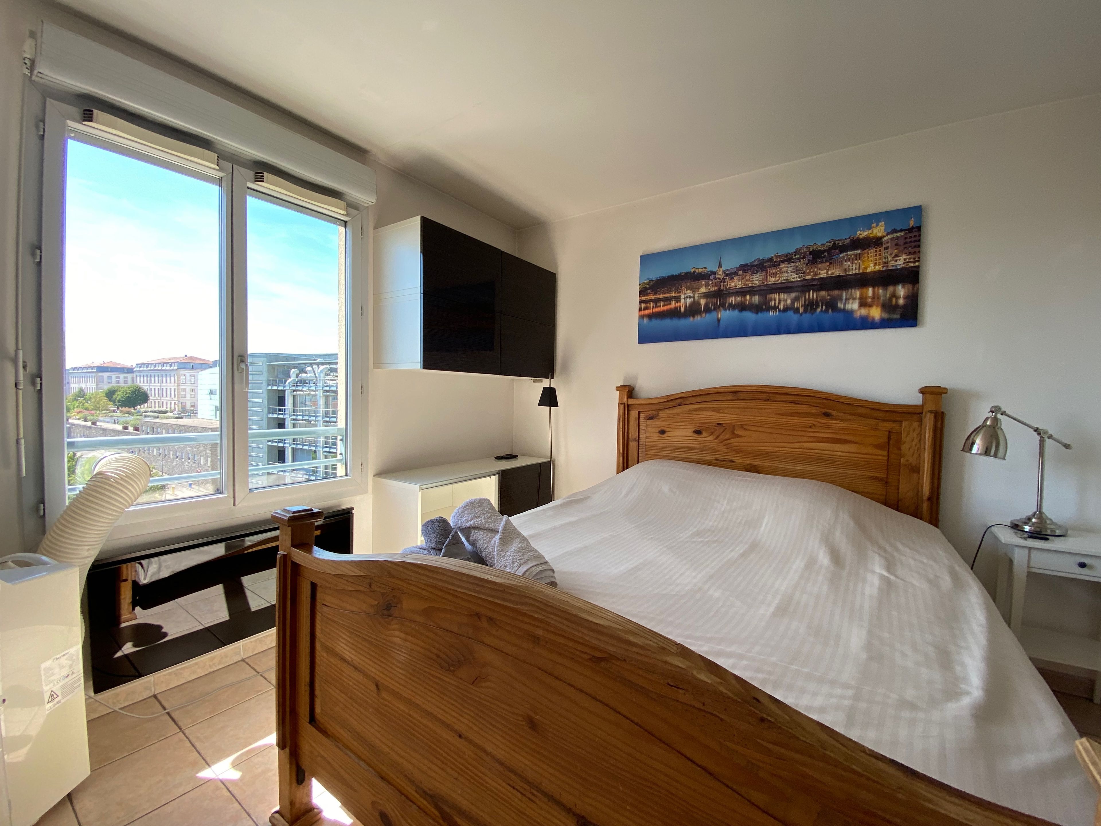 A bedroom with a wooden bed, a desk with a chair, a TV mounted on the wall, and a window with a city view.