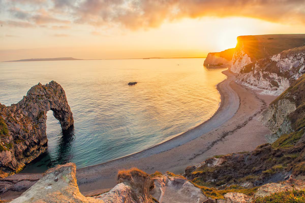 Paisaje costero con una formación rocosa destacada, una costa curva y una puesta de sol.
